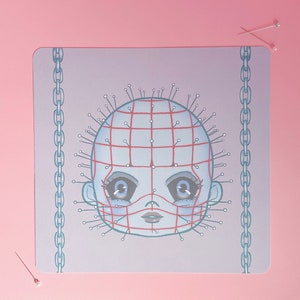 May include: A square, light purple mousepad featuring a stylized illustration of a doll-like face with pins and a red grid pattern. The design includes a chain border and is set against a pink background.