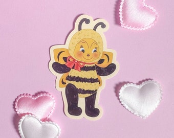May include: A yellow and black bee sticker with a red bow, wearing a yellow and white fuzzy costume. The bee is smiling and has big eyes. The sticker is on a pink background with white and pink hearts.