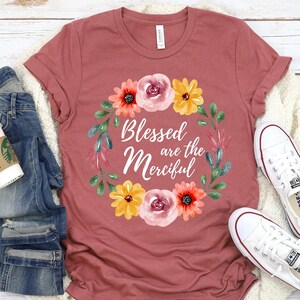 Op de afbeelding: Een oudroze t-shirt met de tekst "Blessed are the Merciful" omringd door een bloemenkrans. De afbeelding bevat een blauwe jeans, een koffiekopje, een crèmekleurige trui en witte Converse sneakers.