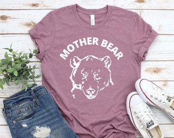 May include: A mauve-colored t-shirt with the words "MOTHER BEAR" above a white bear illustration. The shirt is paired with blue jeans and white sneakers on a light wood surface.