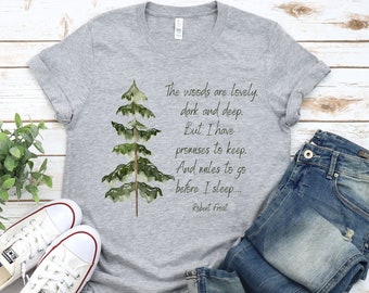 May include: A heather gray t-shirt featuring a watercolor pine tree design and the Robert Frost poem, "The woods are lovely, dark and deep." The shirt is paired with blue jeans and white sneakers.