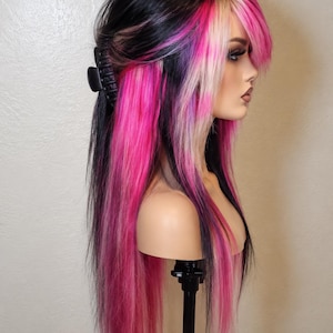 May include: A long wig featuring a layered cut with a mix of black, bright pink, and platinum blonde hair. The hair is styled with a black claw clip securing the top section, while long strands frame the face with prominent pink and blonde highlights.