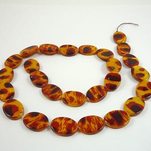 May include: A necklace featuring oval-shaped beads in shades of amber, yellow, and dark brown. The beads have a marbled pattern, strung together with a thin, brown cord. The necklace is displayed on a white background.