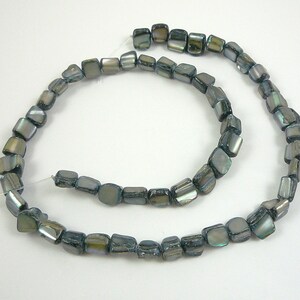 May include: A strand of iridescent, square-shaped beads in shades of gray, green, and blue. The beads are strung together on a thin, white string, creating a curved shape against a white background. The beads have a polished, reflective surface.