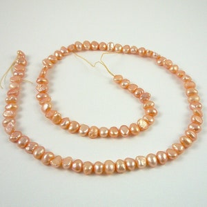 May include: A strand of peach-colored freshwater pearls. The pearls are irregular in shape and have a slightly iridescent sheen.