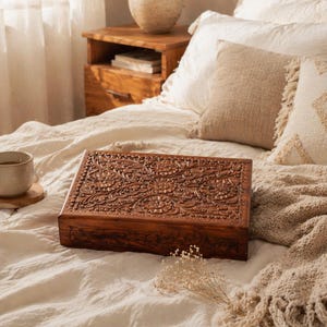 May include: A rectangular, dark brown wooden box with intricate floral carvings on the lid sits on a bed. The box is surrounded by cream-colored bedding, pillows, and a beige throw blanket. A cup of coffee rests on a wooden coaster.