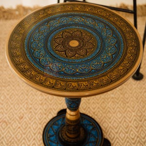 Pode incluir: Uma mesa lateral redonda com um design decorativo pintado à mão. O tampo apresenta um motivo floral central em azul e dourado, rodeado por padrões intrincados. A mesa tem uma base de pedestal e mede aproximadamente 50 cm de diâmetro.