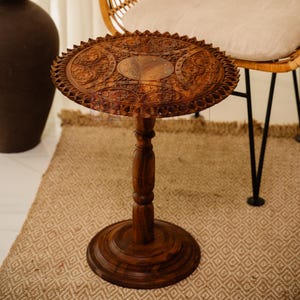 May include: A handcrafted, dark brown wooden side table with intricate carved floral patterns on the round tabletop. The table has a single, turned pedestal base and a decorative edge. It stands on a patterned rug.