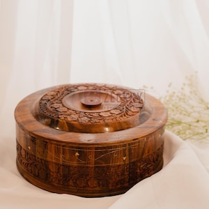 Può includere: Una scatola rotonda in legno con intagli intricati e un coperchio. La scatola è in legno scuro e ha una finitura naturale. Il coperchio ha un piccolo pomo al centro.