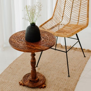 May include: A carved wooden side table with a black vase and white flowers on top. The table has a round top and a single pedestal base. The table is sitting on a woven rug in front of a wicker chair.