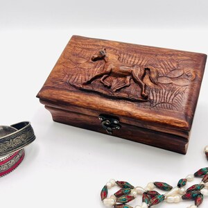 May include: A wooden jewelry box with a carved horse design on the lid. The box is closed and has a brass latch. The box is sitting on a white surface next to a pearl and turquoise beaded necklace and two silver bracelets.