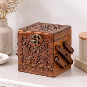 Op de afbeelding: Een vierkante houten doos met ingewikkelde bloemensnijwerk en een scharnierend deksel. De doos is gebeitst in een warmbruine kleur en heeft een metalen sluiting. Op een witte ondergrond.