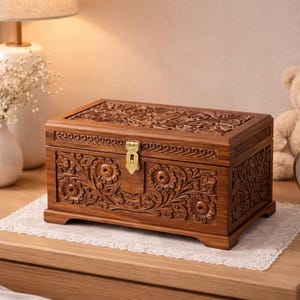 Puede incluir: Un joyero de madera marrón oscuro con intrincadas tallas florales. La caja es rectangular, con tapa con bisagras y un cierre dorado. Descansa sobre un tapete de encaje blanco. Una lámpara, flores y un oso de peluche están en el fondo.