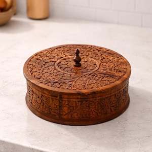 Puede incluir: Una caja redonda de madera con intrincadas tallas florales. La caja es de color marrón cálido y presenta un pomo decorativo en la tapa. Las tallas detalladas cubren la tapa y los lados, creando una superficie texturizada.