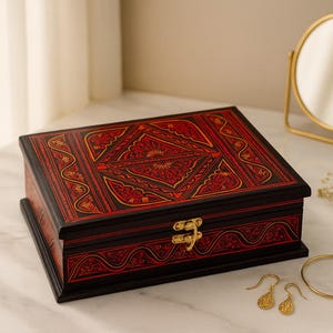 Pode incluir: Uma caixa de madeira decorativa com um padrão vermelho e dourado detalhado sobre um fundo preto. A caixa é retangular com um fecho dourado. Brincos dourados, uma pulseira e um pequeno espelho dourado também são visíveis.