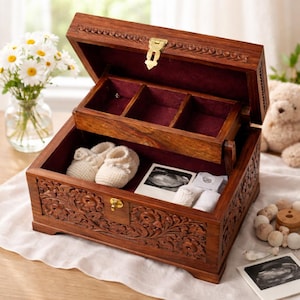 May include: Open wooden keepsake box with carved floral details, a brass clasp, and a burgundy velvet interior. Inside are baby booties, ultrasound photos, and small white items. The box is open, revealing a smaller tray with compartments.