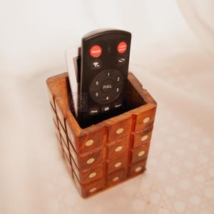 May include: A wooden remote control holder with brass accents. The holder has a square shape and is filled with two remote controls. One remote is black and the other is white.