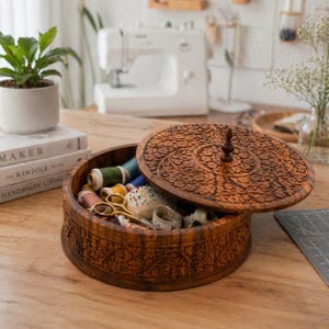 May include: A round, wooden sewing box with an open lid, revealing spools of thread, scissors, and a pincushion. The box and lid are intricately carved with floral patterns. The box is on a wooden table, with a sewing machine in the background.