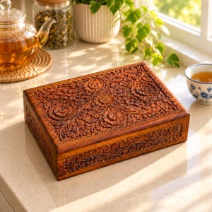 Puede incluir: Una caja rectangular de madera con intrincados patrones florales tallados a mano que cubren toda la superficie superior y los lados. La madera tiene un acabado cálido en tono marrón medio con detalles oscuros en el relieve.