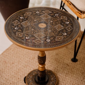 May include: A round side table with an ornate, dark brown and gold patterned top. The table has a gold-colored pedestal base and a decorative, carved central support. The table sits on a patterned beige rug.