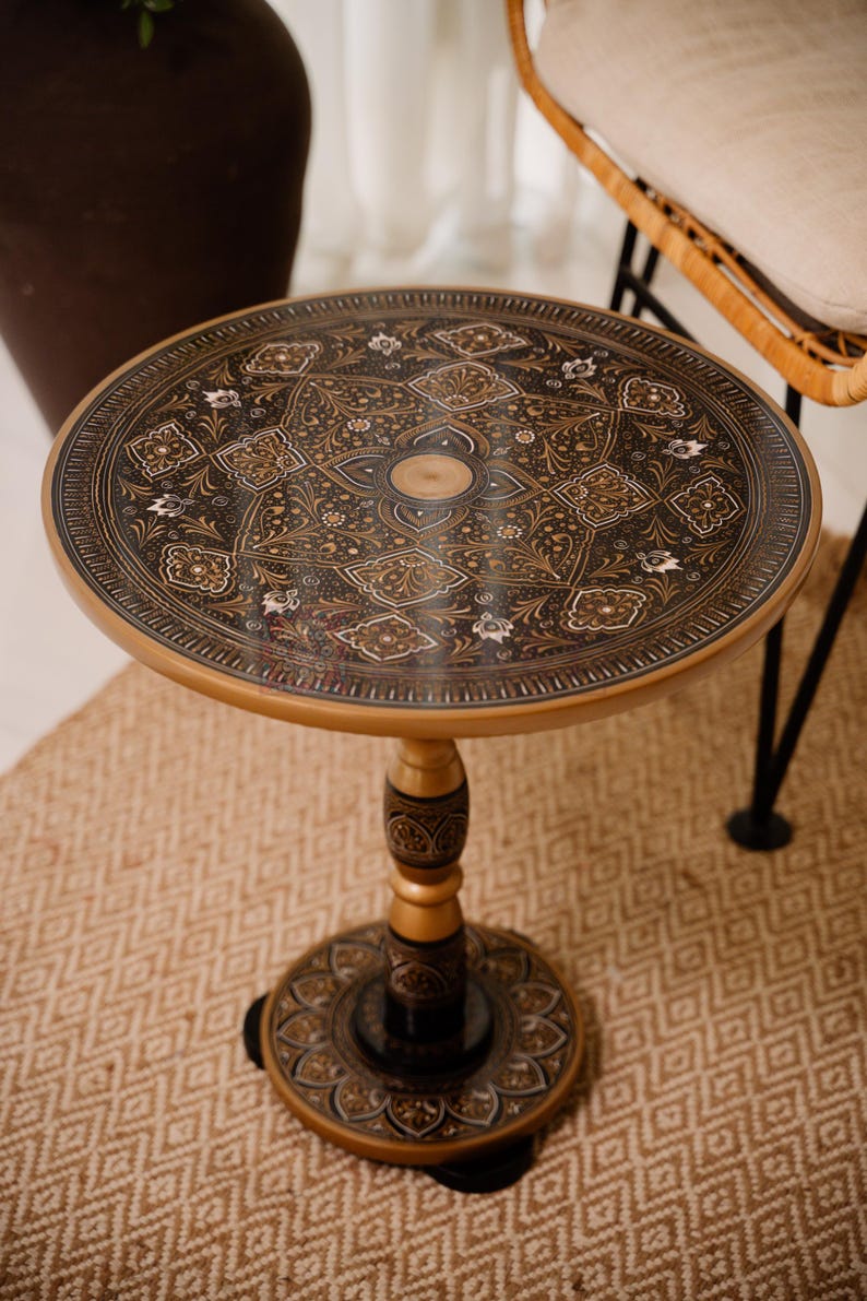 May include: A round side table with an ornate, dark brown and gold patterned top. The table has a gold-colored pedestal base and a decorative, carved central support. The table sits on a patterned beige rug.