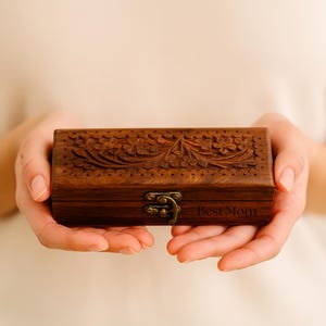 Puede incluir: Una caja de madera rectangular con intrincadas tallas florales en la tapa. La caja es de color marrón cálido y tiene un cierre de metal. Las palabras "Best Mom" están grabadas en el lateral. La caja se sostiene con dos manos.