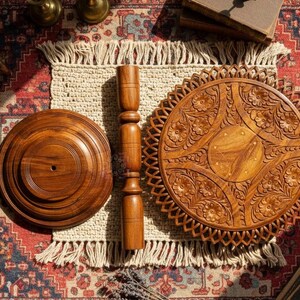 May include: A disassembled wooden table with a round base, a central support, and a decorative top. The table components are made of a warm, medium-toned wood and are arranged on a patterned rug with a fringed placemat.