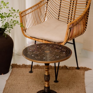 May include: A round, decorative side table with an ornate, dark metal top and a gold-colored pedestal base. The table is approximately 20 inches in diameter and stands on three black legs. A woven rattan chair with a cream-colored cushion is in the background.