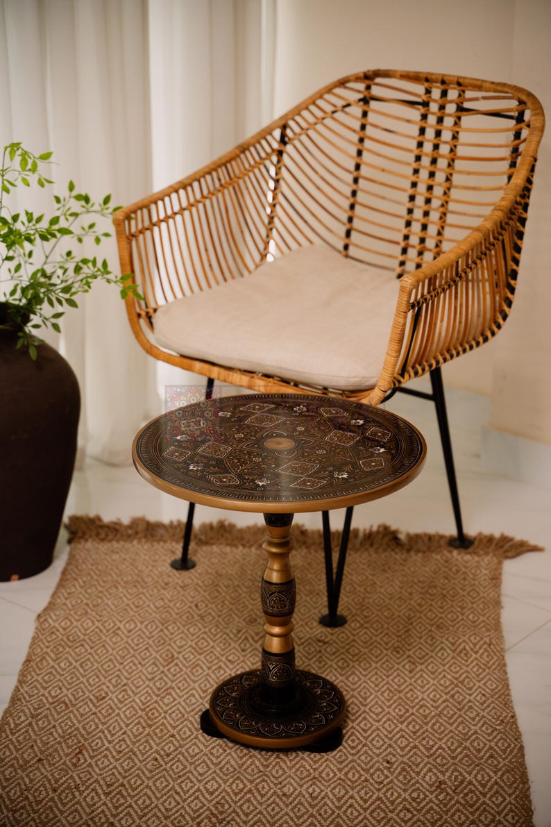 May include: A round, decorative side table with an ornate, dark metal top and a gold-colored pedestal base. The table is approximately 20 inches in diameter and stands on three black legs. A woven rattan chair with a cream-colored cushion is in the background.