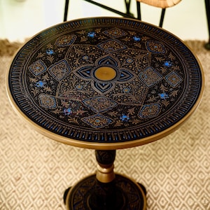 May include: Round black side table with intricate gold and blue floral patterns. The table has a gold rim and a pedestal base. The design is symmetrical and detailed, creating an elegant aesthetic. The table is a decorative furniture piece.