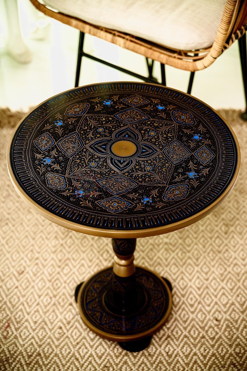 May include: Round black side table with intricate gold and blue floral patterns. The table has a gold rim and a pedestal base. The design is symmetrical and detailed, creating an elegant aesthetic. The table is a decorative furniture piece.