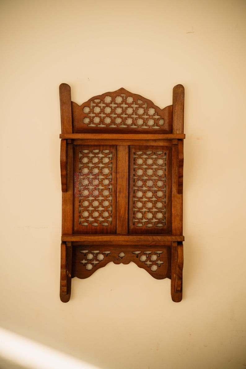 May include: A brown wooden wall shelf featuring two horizontal tiers and a central section with two vertical panels. The structure is adorned with intricate, repeating geometric lattice cutouts throughout the top, middle, and bottom sections.