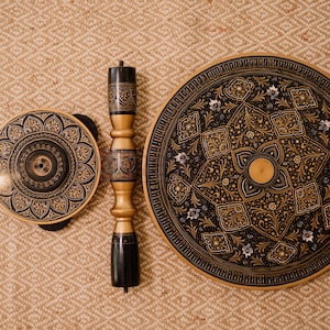 May include: A set of three wooden decorative pieces with intricate black and gold floral patterns. The set includes two circular tabletops and a central support column. The pieces are displayed on a textured, neutral-toned surface.