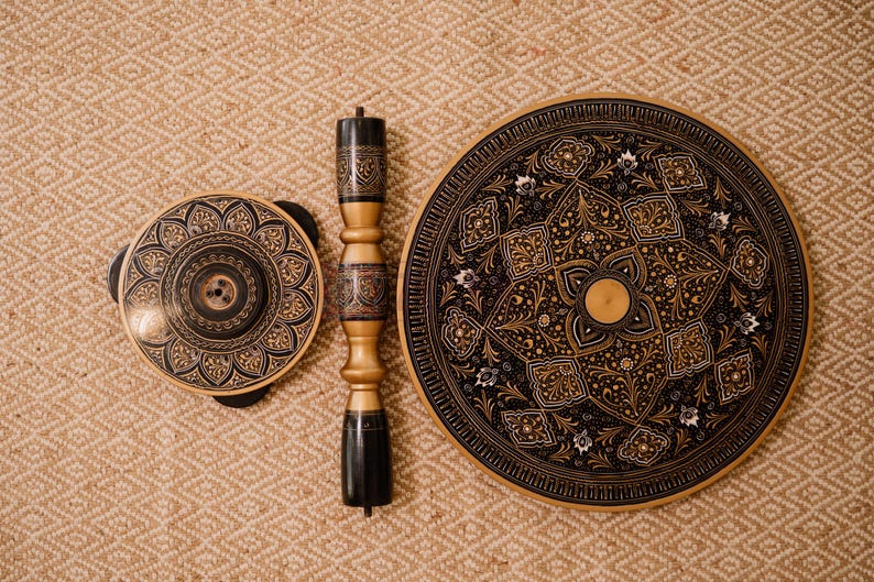 May include: A set of three wooden decorative pieces with intricate black and gold floral patterns. The set includes two circular tabletops and a central support column. The pieces are displayed on a textured, neutral-toned surface.
