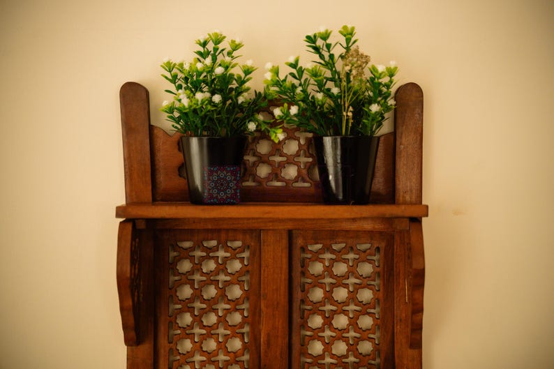 May include: A brown wooden wall shelf featuring intricate lattice cutouts and two black pots containing artificial green plants with small white flowers. The left pot displays a small square sticker with a blue and purple geometric pattern.