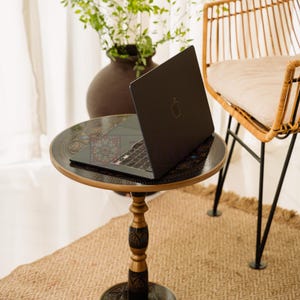 May include: A black laptop with the Apple logo sits on a small, round table with an ornate, dark-colored top and a decorative pedestal base. The table is on a woven rug, with a wicker chair and a vase of greenery in the background.
