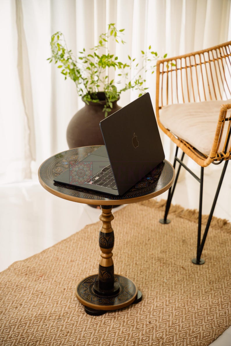 May include: A black laptop with the Apple logo sits on a small, round table with an ornate, dark-colored top and a decorative pedestal base. The table is on a woven rug, with a wicker chair and a vase of greenery in the background.