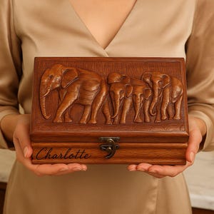 May include: A rectangular wooden box with a carved relief of three elephants. The box is a warm brown color and has a metal clasp. The name "Charlotte" is engraved on the front.