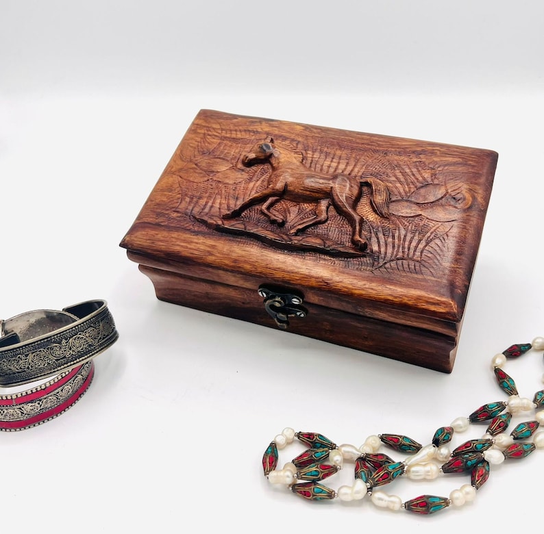 May include: A wooden jewelry box with a carved horse design on the lid. The box is closed and has a brass latch. The box is sitting on a white surface next to a pearl and turquoise beaded necklace and two silver bracelets.