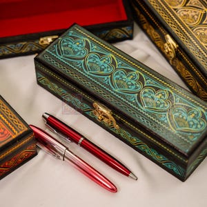 May include: Several rectangular wooden boxes with intricate designs and a pair of pens. The boxes feature ornate patterns in teal, gold, and orange, with heart and floral motifs. The pens are red and silver.