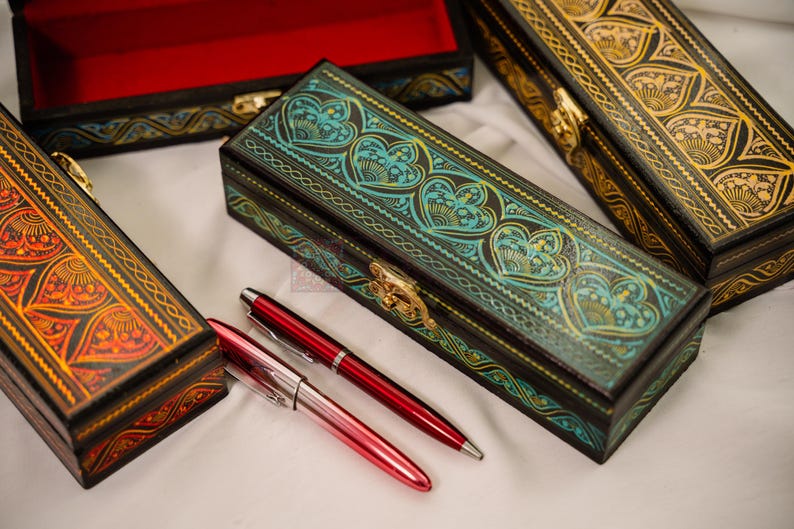 May include: Several rectangular wooden boxes with intricate designs and a pair of pens. The boxes feature ornate patterns in teal, gold, and orange, with heart and floral motifs. The pens are red and silver.