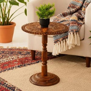 May include: A small, round, wooden side table with intricate carved detailing on the tabletop. The table is a warm brown color and has a single central leg. A small potted plant sits on top of the table. The table is next to a cream-colored armchair.