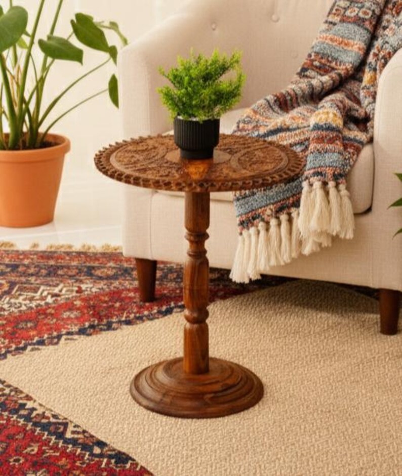 May include: A small, round, wooden side table with intricate carved detailing on the tabletop. The table is a warm brown color and has a single central leg. A small potted plant sits on top of the table. The table is next to a cream-colored armchair.