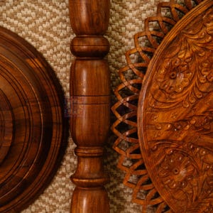 May include: Close-up of a wooden decorative stand, featuring a round base, a central pillar, and a carved floral design on the tabletop. The wood has a rich brown color and intricate details, showcasing craftsmanship.