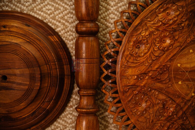 May include: Close-up of a wooden decorative stand, featuring a round base, a central pillar, and a carved floral design on the tabletop. The wood has a rich brown color and intricate details, showcasing craftsmanship.