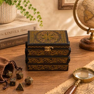 May include: A rectangular black wooden box with gold-colored intricate patterns on the lid and sides, featuring a metal latch. Beside the box are several multi-sided dice in metallic gold and black, along with a brown fabric drawstring pouch.