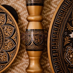 May include: Close-up of decorative wooden objects with intricate black and gold designs. The image features a central, ornate pillar and two circular plates with detailed patterns, showcasing a blend of craftsmanship and artistry.
