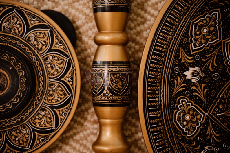 May include: Close-up of decorative wooden objects with intricate black and gold designs. The image features a central, ornate pillar and two circular plates with detailed patterns, showcasing a blend of craftsmanship and artistry.