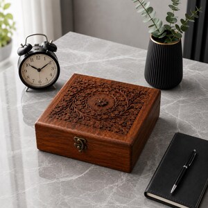 May include: A dark wooden box with intricate floral carvings on the lid, secured with a metal clasp. A black alarm clock, a black vase with gold trim, and a black notebook with a pen are also on the gray surface.