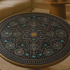 May include: Round coffee table with a black surface featuring an intricate floral design in gold and teal. The table has a gold-colored metal frame and a decorative edge. A stylish piece for living rooms.